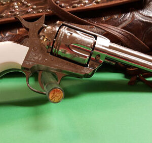 Close-up of a shiny revolver with a white grip on a green surface, with brown leather straps in the background.